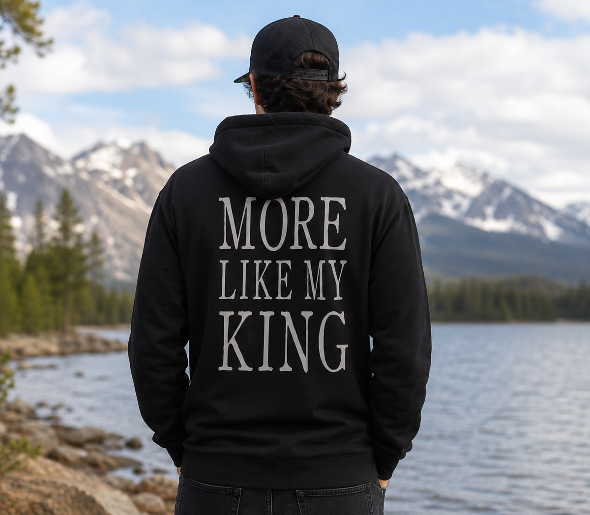 A man viewed from behind wearing a black hoodie with the phrase 'MORE LIKE MY KING' printed in large white serif letters on the back, standing outdoors in front of a lake and snow-capped mountains.