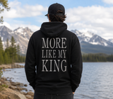 A man viewed from behind wearing a black hoodie with the phrase 'MORE LIKE MY KING' printed in large white serif letters on the back, standing outdoors in front of a lake and snow-capped mountains.