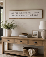 A rustic framed wall sign with the text 'AS FOR ME AND MY HOUSE WE WILL SERVE THE LORD' hangs above a wooden console table in a styled home entryway. The table holds a potted plant, books, a bowl, a small picture frame, and a table lamp, with a woven basket and throw blanket underneath.