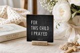 “For This Child I Prayed” 4 x 4-inch shelf sitter in the white lettering on black distressed finish, displayed on a nursery window ledge.