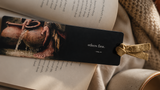 A long black book bookmark with a gold ribbon loop, featuring the text 'others first.' and an artistic print of clay pots, resting on open books with a cozy beige blanket and ceramic mug in the background.
