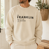 A man wearing a cream-colored crewneck sweatshirt with the text 'IN FRANKLIN AS IT IS IN HEAVEN' printed in black on the front, paired with matching cream sweatpants and a beige baseball cap, standing in a bright, modern indoor setting.