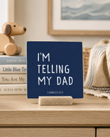 A navy blue square sign with white text reading “I'M TELLING MY DAD” and small text “2 CHARITIES 2015,” displayed on a wooden stand on a light wood surface in a styled room setting.