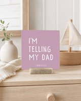 A square lavender card with white text reading 'I'M TELLING MY DAD' and 'PROVERBIALIST JESUS' below, displayed on a wooden stand on a light wood dresser with home decor props.