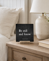 Be Still and Know 4x4 Christian Shelf Sitter on a light wood nightstand with a speckled lamp and soft cream bedding.