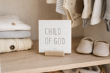 “Child of God” 4 x 4-inch shelf sitter in the gray lettering on white finish, displayed on a nursery closet shelf.