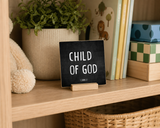 Close-up of “Child of God,” a 4 x 4-inch shelf sitter in the white lettering on black distressed finish in a family room built-in.