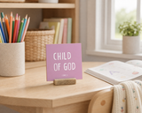 Close-up of “Child of God,” a 4 x 4-inch shelf sitter in the white lettering on pink finish on a homeschool desk.