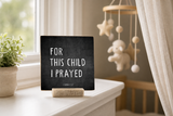 “For This Child I Prayed” 4 x 4-inch shelf sitter in the white lettering on black distressed finish, displayed on a nursery window ledge.