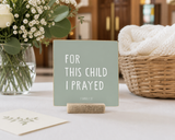 “For This Child I Prayed,” a 4 x 4-inch shelf sitter in the white lettering on green finish, styled on a baby dedication table.