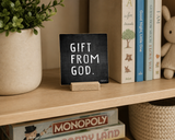 “Gift from God” 4 x 4-inch shelf sitter in the white text on black distressed finish, displayed in a family room built-in.