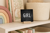 “Girl” 4 x 4-inch shelf sitter in the white lettering on black distressed finish, displayed on a rainbow nursery bookcase.