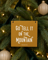 Gold 4x4 Go Tell It on the Mountain ornament with metal wire hanger on a noble fir with deep green velvet ribbon, cream baubles, and warm lights.