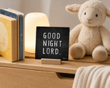 “Good Night Lord” 4 x 4-inch shelf sitter in the white text on black distressed finish, displayed in a toddler bedside crate.