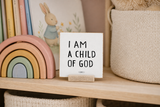 “I Am a Child of God,” a 4 x 4-inch shelf sitter in the black lettering on white distressed finish, styled on a rainbow nursery bookcase.