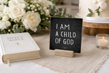 “I Am a Child of God” 4 x 4-inch shelf sitter in the white lettering on black distressed finish, displayed on a baptism celebration table.