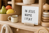 “In Jesus' Name I Play,” a 4 x 4-inch shelf sitter in the black letters with white distressed finish, styled on a play kitchen shelf.