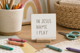 Close-up of “In Jesus' Name I Play,” a 4 x 4-inch shelf sitter in the gray letters with white distressed finish on a kids craft table.