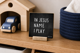“In Jesus' Name I Play” 4 x 4-inch shelf sitter in the white letters with black distressed finish, displayed on a toy car shelf.