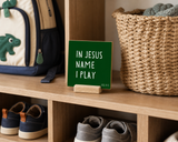 “In Jesus' Name I Play,” a 4 x 4-inch shelf sitter in the white letters with green finish, styled on a family entryway cubby.