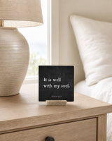 It Is Well With My Soul 4x4 Christian Shelf Sitter on a light nightstand with a ceramic lamp and soft white bedding.