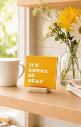 It's Gonna Be Okay 4x4 Christian Shelf Sitter on a sunlit kitchen shelf with a white mug and cheerful yellow flowers.
