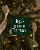 Dark Green 4x4 Jesus Is Coming to Town ornament with metal wire hanger on a noble fir with small brass stars, cream ribbon, and warm lights.