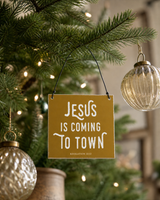 Gold 4x4 Jesus Is Coming to Town ornament with metal wire hanger on a noble fir with champagne ornaments and warm lights.