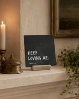 Keep Loving Me 4x4 Christian Shelf Sitter on a plaster mantel with a lit candle, brass vessel, and trailing greenery.
