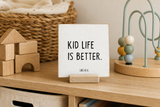 “Kid Life Is Better,” a 4 x 4-inch shelf sitter in the black lettering on white distressed finish, styled on a playroom cubby.