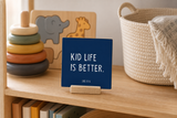 Close-up of “Kid Life Is Better,” a 4 x 4-inch shelf sitter in the white lettering on blue finish on a montessori play shelf.