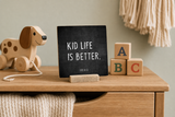 “Kid Life Is Better” 4 x 4-inch shelf sitter in the white lettering on black distressed finish, displayed in a toy chest display.