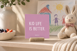 “Kid Life Is Better,” a 4 x 4-inch shelf sitter in the white lettering on pink finish, styled in a family breakfast nook.