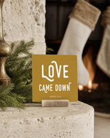 Gold 4x4 Love Came Down Christmas Shelf Sitter in a stone fireplace hearth with its rustic wood slot stand.