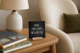 “May the Lord Be with You” 4 x 4-inch shelf sitter in the white lettering on black distressed finish, displayed on a kids reading nook side table.