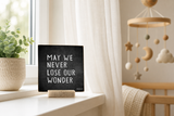 “May We Never Lose Our Wonder” 4 x 4-inch shelf sitter in the white lettering on black distressed finish, displayed on a nursery window ledge.