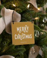 Gold 4x4 Merry Christmas ornament with metal wire hanger on a Fraser fir with clear glass drops, linen ribbon, and candlelike lights.