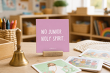 “No Junior Holy Spirit,” a 4 x 4-inch shelf sitter in the white letter on pink finish, styled on a preschool teacher desk.