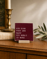 Our Sins They Are Many, His Mercy Is More 4x4 Christian Shelf Sitter on a dark wood console with a brass candlestick and greenery.