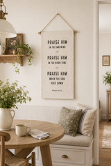 A vertical fabric wall hanging with black typographic text reading 'PRAISE HIM IN THE MORNING, PRAISE HIM IN THE MOON TIME, PRAISE HIM WHEN THE SUN GOES DOWN' hangs on a cream wall above a cushioned bench in a styled interior room.
