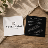 Two square Tetelestai prayer cards displayed on a rustic wooden surface with a linen cloth, leafy sprig, and ceramic bowl as styled props. One card is white with black text/logo, and the other is black with white text.
