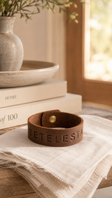 A brown leather cuff bracelet with the name 'TETELESTAI' embossed on the front, resting on a white linen cloth on a wooden table. In the background, a stack of books and a ceramic vase are visible in a warm, softly lit interior setting.