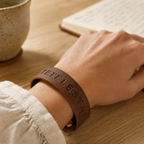 A close-up of a person's hand resting on an open book on a wooden table, wearing a brown leather wristband engraved with 'JETEL ESTAI'.