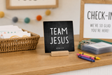 “Team Jesus” 4 x 4-inch shelf sitter in the white lettering on black distressed finish, displayed in a sunday school check-in.