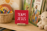 Close-up of “Team Jesus,” a 4 x 4-inch shelf sitter in the white lettering on red finish on a children's church shelf.