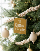 Gold 4x4 The Kingdom of God Is Here ornament with metal wire hanger on a Christmas tree with woven cream garland and pale wood ornaments.