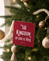 Red 4x4 The Kingdom of God Is Here ornament with metal wire hanger on a tree with woven cream garland, pale wood ornaments, and aged brass.