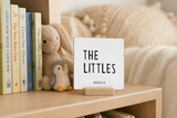 “The Littles,” a 4 x 4-inch shelf sitter in the black lettering on white distressed finish, styled on a sibling bookshelf.