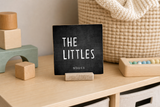 “The Littles” 4 x 4-inch shelf sitter in the white lettering on black distressed finish, displayed on a playroom cubby.