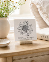 All Things Beautiful 4x4 Christian Shelf Sitter with floral artwork on a light bedside table beside daisies and neutral pottery.